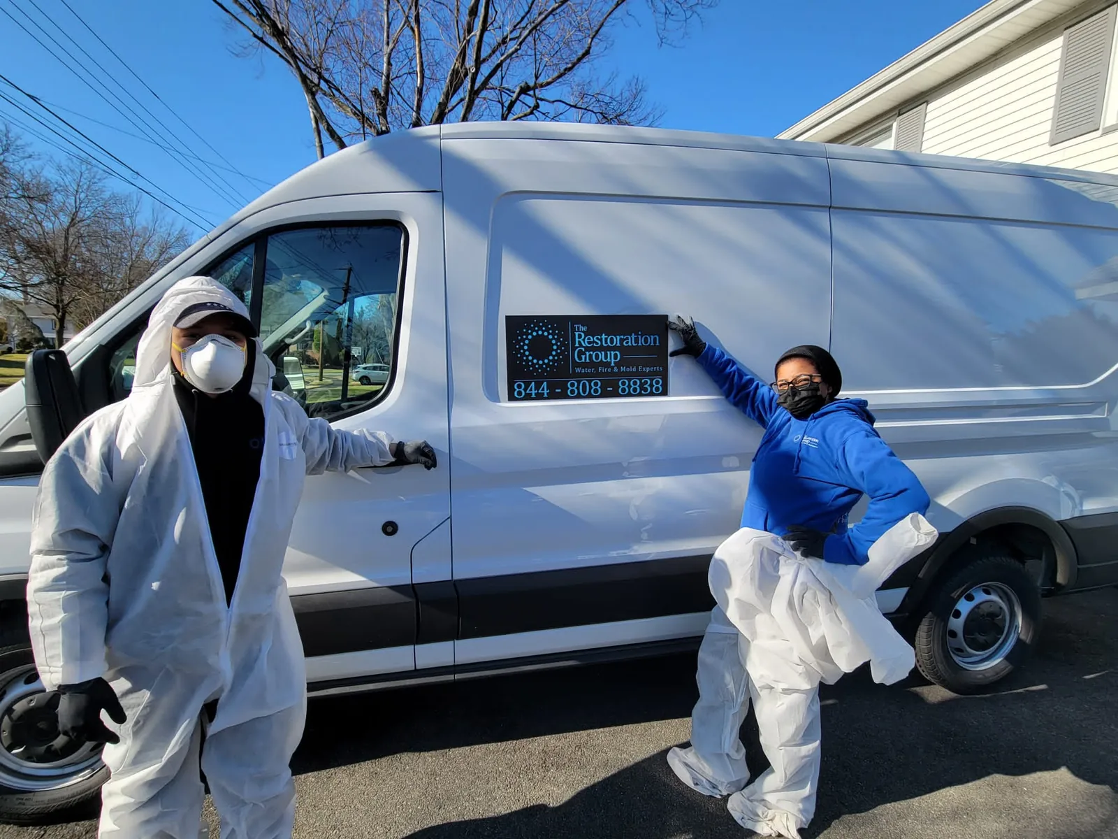 Two The Restoration Group technicians in protective suits beside a lettered company van on a New Jersey street