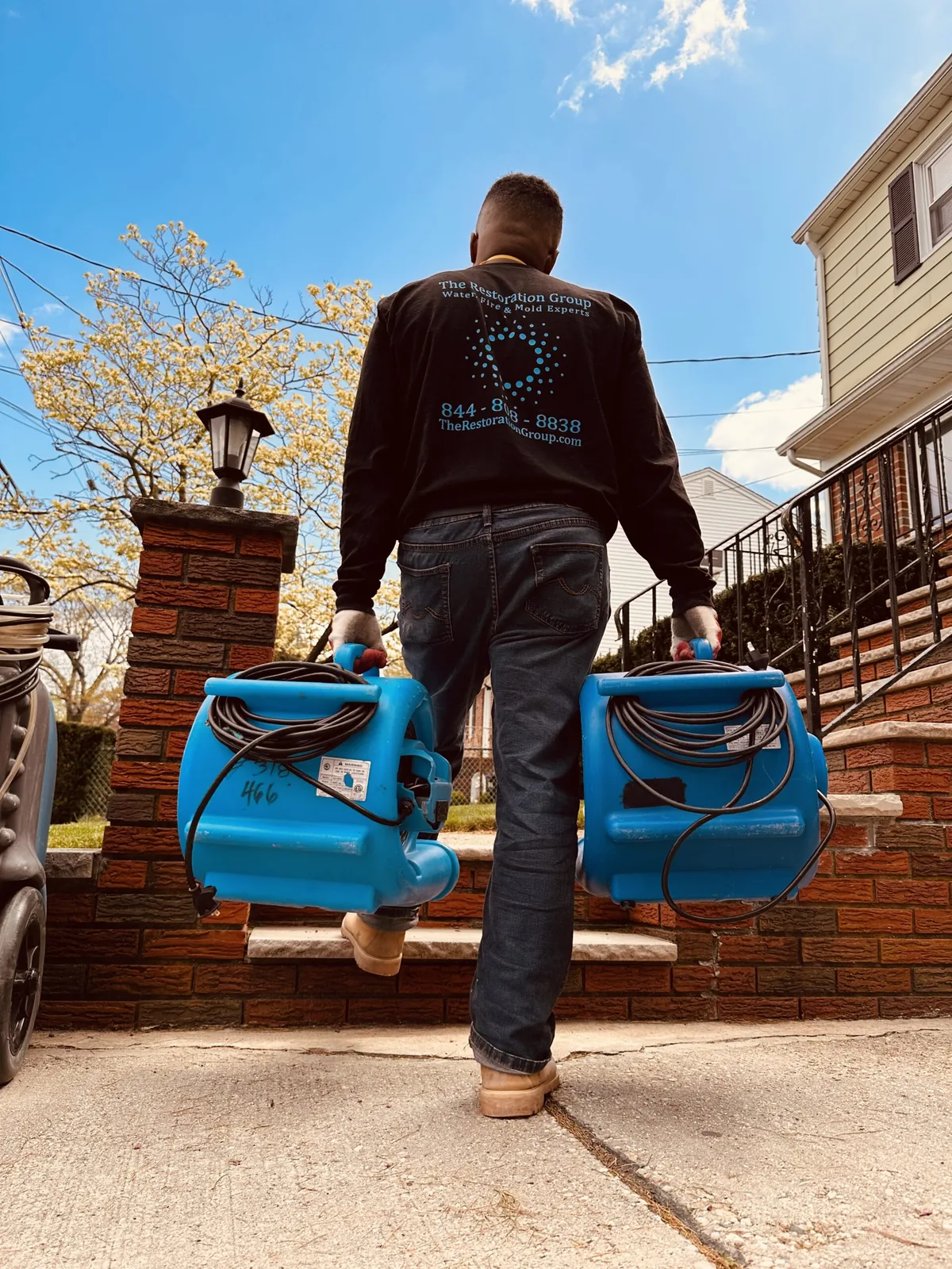 Technician in a The Restoration Group shirt carrying two air movers up the front steps of a home